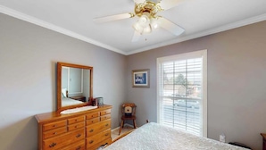 A bedroom with a wooden dresser, a bed with a patterned bedspread, a ceiling fan, a window with blinds, and a framed picture on the wall. - Springs Sanctuary - Dog-Friendly Home • Decks & Fireplaces (Colorado Springs)