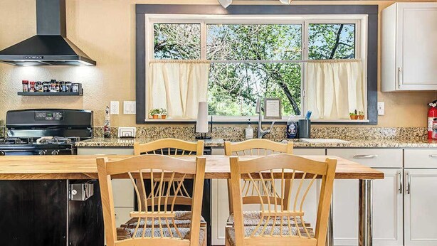 A kitchen with a dining table, chairs, a stove, a range hood, and a window with curtains. - Rocky Mountain Rustic - Mountain-View Home • AC & Fast WiFi (Colorado Springs)