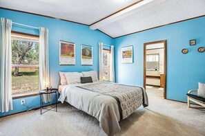 A bedroom with a large bed, a bedside table, a window with curtains, and artwork on the wall. - Rocky Mountain Rustic - Mountain-View Home • AC & Fast WiFi (Colorado Springs)