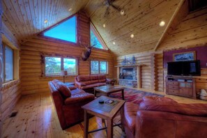 A log cabin interior with a fireplace, leather sofas, a television, and a mounted deer head. - Black Diamond Lodge - Log Lodge • Hot Tub & Game Room (Lead)