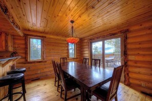 Ein Interieur einer Holzhütte mit Esstisch, Stühlen, großen Fenstern und einer Hängeleuchte.