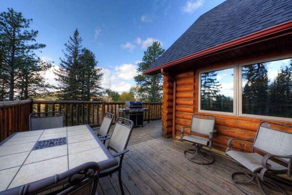 Eine Holzterrasse mit Tisch und Stühlen, ein Grill und eine Hütte mit großen Fenstern.