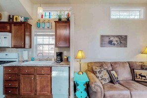 A kitchen with wooden cabinets, a dishwasher, and a window. There's a couch with patterned cushions and a lamp. - Cub Cottage - Cozy Tiny Home • Mtn View, Dogs OK (Woodland Park)