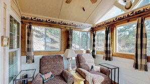 Interior acolhedor de uma cabana com duas poltronas estampadas, uma mesinha e um abajur. Grandes janelas com cortinas oferecem vista da floresta ao redor.