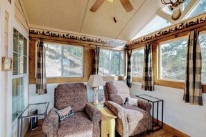 Ein gemütliches Blockhaus-Interieur mit zwei gemusterten Sesseln, einem kleinen Tisch und einer Lampe. Große Fenster mit Vorhängen bieten Blick auf den umliegenden Wald.