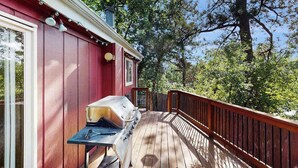 Une terrasse en bois avec un barbecue, entourée d’arbres et bordée d’un bâtiment rouge.