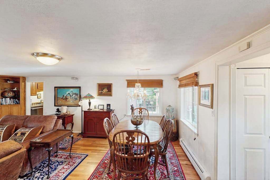A dining area with a wooden table and chairs, a red and blue patterned rug, and a view of the outdoors through a window.