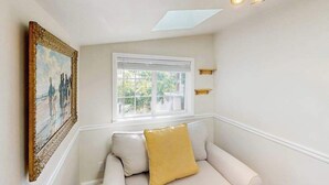 Une pièce avec un canapé blanc, un tableau au mur, une fenêtre avec des stores et un luminaire au plafond.