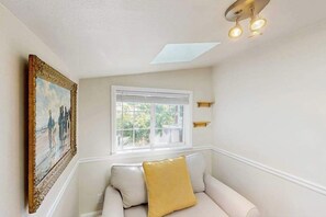 Ein Zimmer mit einem weißen Sofa, einem Gemälde an der Wand, einem Fenster mit Jalousien und einer Deckenleuchte.