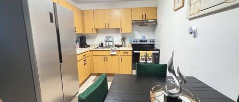 A compact kitchen with wooden cabinets, a stainless steel refrigerator, a dining table with a green cushion, and a decorative metal sculpture.