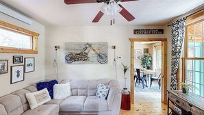A cozy living room with a ceiling fan, a couch with decorative pillows, a motorcycle artwork on the wall, and a sign that reads 'Family & Friends Gather Here'. - Mountain View Escape - Dog-Friendly Cabin • Amazing Views (Lead)