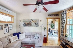 Ein gemütliches Wohnzimmer mit Deckenventilator, einer Couch mit Zierkissen, einem Motorradbild an der Wand und einem Schild, auf dem steht 'Familie & Freunde versammeln sich hier'.