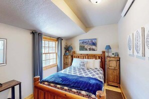 A bedroom with a wooden bed, a window with curtains, a painting on the wall, and a small cabinet. - Mountain View Escape - Dog-Friendly Cabin • Amazing Views (Lead)
