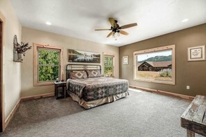 A bedroom with a bed, a ceiling fan, two windows, and a view of a house. - Klassy Lodge - New Lodge • Private Hot Tub & Game Room (Sturgis)