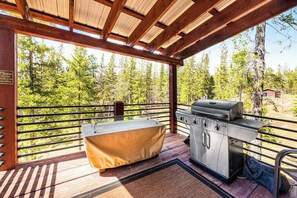 Eine Terrasse im Freien mit Grill und überdachtem Tisch, umgeben von einer Holzdielenveranda und einem Metallgeländer, mit Blick auf einen bewaldeten Bereich.