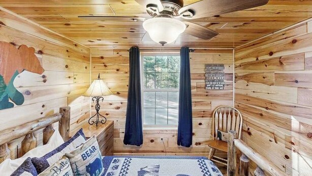 A cabin interior with wooden walls, a ceiling fan, a bed with a blue and white patterned bedspread, a chair, and a window with curtains. - Meandering Moose Cabin - Secluded Wood Cabin w/ Big Deck (Florissant)