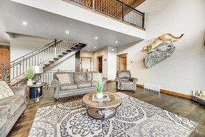 Ein geräumiges Wohnzimmer mit einer großen Treppe, Ledersofas, einem gemusterten Teppich und einer an der Wand befestigten Katzenattrappe.