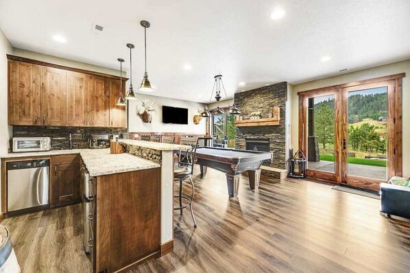 A modern kitchen with wooden cabinets, a central island, and a pool table. - Cedar Lodge on 17- Hot Tub Home Near Mt Rushmore (Sturgis)