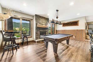 A pool table in a room with a stone fireplace, wooden flooring, and a deer head decoration. - Cedar Lodge on 17- Hot Tub Home Near Mt Rushmore (Sturgis)