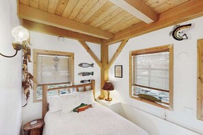 Ein Schlafzimmer mit Holzbalkendecke, einem Bett mit weißer Bettwäsche, zwei Fenstern mit Jalousien und Fischdekorationen an der Wand.