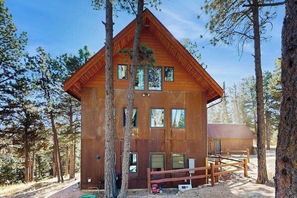 A two-story wooden cabin with a fenced yard, surrounded by trees. - Treehouse Retreat • Pikes Peak Views with Hot Tub (Florissant)