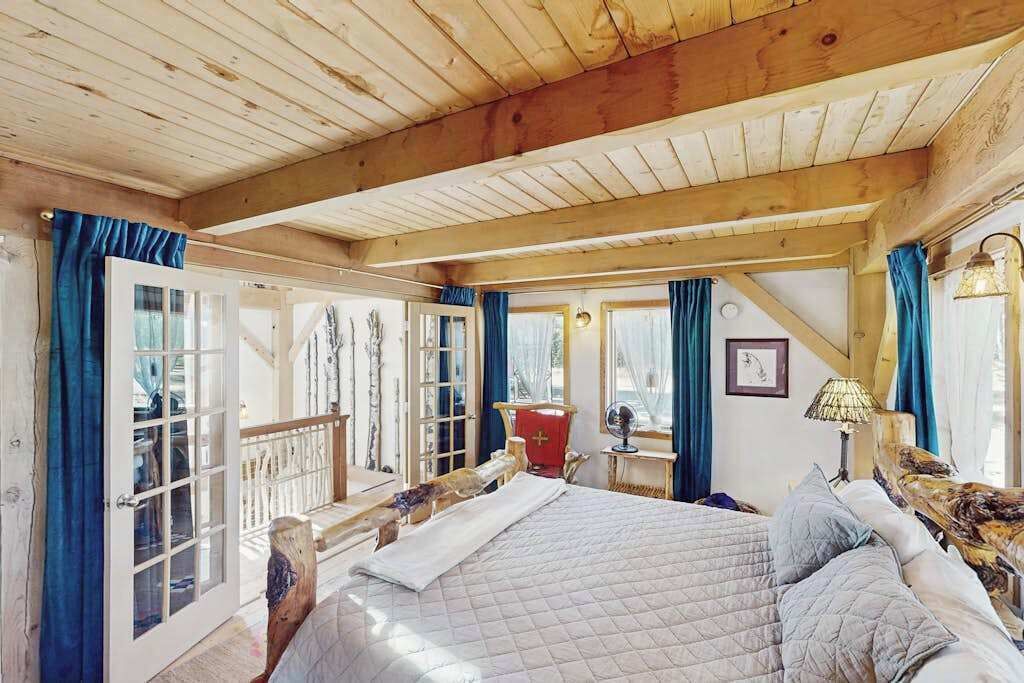 A bedroom with a large bed, wooden beams, a balcony, and a view of the outdoors.