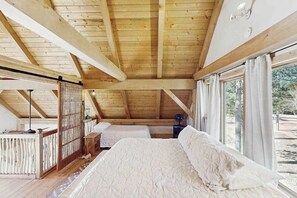A bedroom with a wooden ceiling, a bed with white bedding, and a view of the outdoors through large windows.