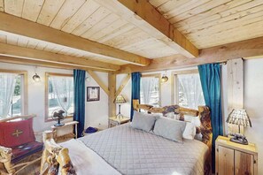 A wooden cabin bedroom with a bed, a wooden headboard, a nightstand, a red chair, and a window with curtains.