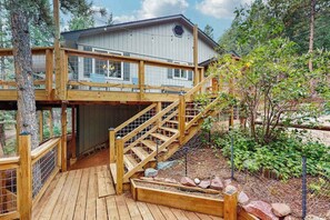 Une terrasse en bois à deux niveaux, avec un escalier menant à une zone abritée, le tout entouré d’arbres et d’un jardin.