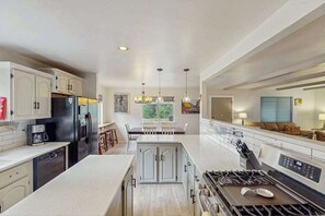 A modern kitchen with white cabinetry, a central island, a built-in stove, and a dining area with a view of the outdoors. - The Lookout at Woodland Park - Dog-Friendly Mtn Home • Fenced Yard (Woodland Park)