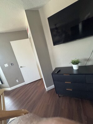 Ein Zimmer mit einem an der Wand befestigten Flachbildfernseher, einer dunkelbraunen Kommode mit Blumentopf und einer Tür mit Lichtschalter.