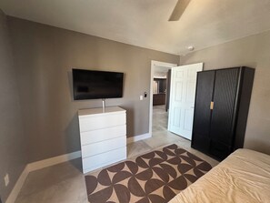 Ein Schlafzimmer mit einem Bett, einem Schminktisch, einem an der Wand befestigten Fernseher und einer Tür zu einem anderen Raum.