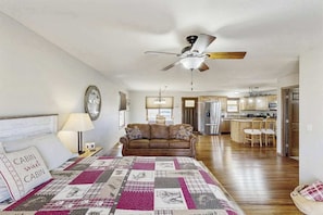 A bedroom with a bed, a nightstand, a lamp, a ceiling fan, a couch, and a view into the kitchen. - Stratobowl Escape - Dual-Unit Home Near Black Hills Fun (Rapid City)