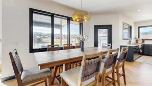 A dining area with a large table and chairs, a hanging light fixture, and large windows offering a view of the city. - The 19th Hole - Golf Simulator Home • Hot Tub & Views (Sturgis)