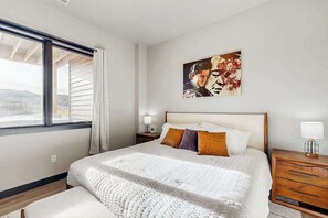 A bedroom with a large bed, a nightstand, a wall-mounted painting, and a window with curtains. - The 19th Hole - Golf Simulator Home • Hot Tub & Views (Sturgis)