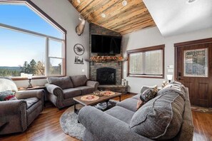 A cozy living room with a fireplace, large windows, and a high ceiling.