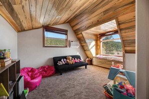 Un dormitorio acogedor en el ático, con techo inclinado, sofá negro, sillón de peluche y ventana con vistas al exterior.