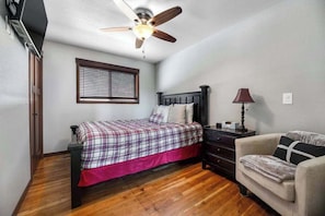 A bedroom with a bed, a ceiling fan, a nightstand, a lamp, a chair, and a window with blinds.