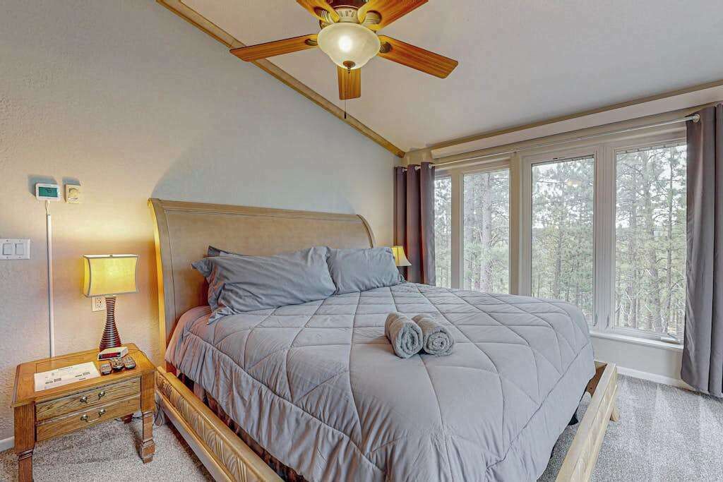 A bedroom with a bed, a nightstand, a lamp, a ceiling fan, and large windows.