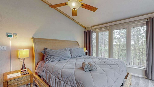 A bedroom with a bed, a nightstand, a lamp, a ceiling fan, and large windows. - Boxelder Bluff - Secluded Home • Mountain Views (Rapid City)