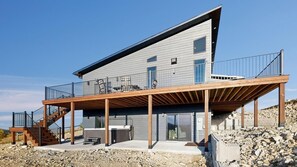 Een modern huis met twee verdiepingen, een houten terras, zitplaatsen buiten en een trap naar de bovenverdieping.