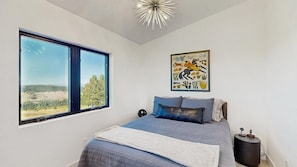 A bedroom with a large window, a bed with blue bedding, a modern light fixture, and a wall-mounted artwork. - Cowgirl Heaven - 360 Views - Luxury Lodge Sleeps 20 (Sturgis)