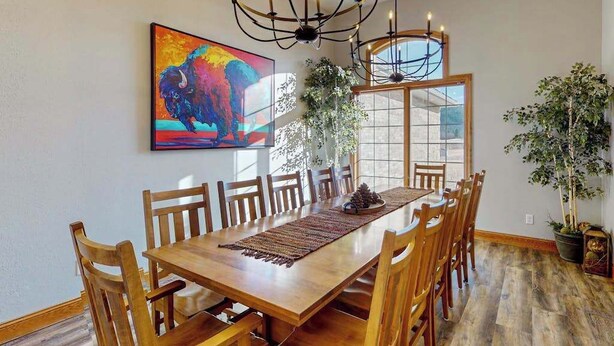 Uma sala de jantar com mesa e cadeiras de madeira, um lustre, um quadro colorido e uma janela grande.
