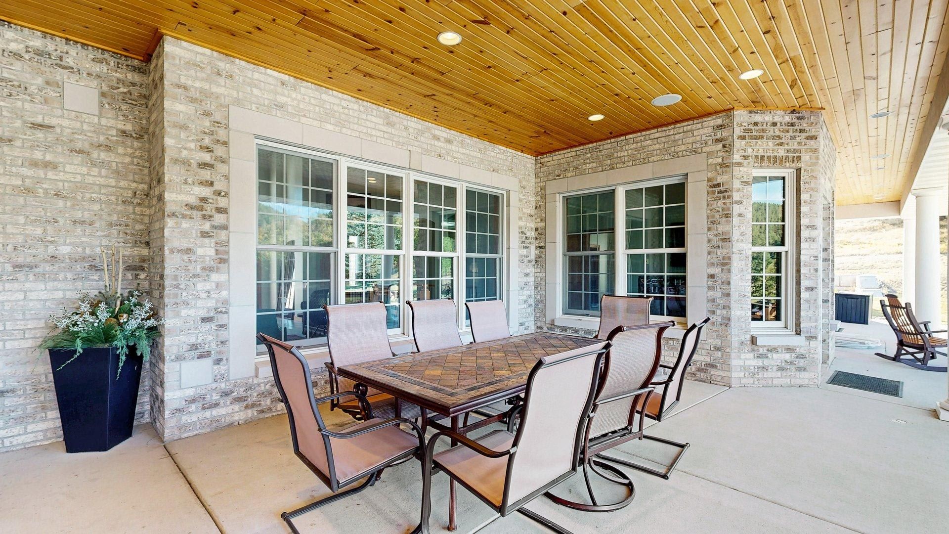 An outdoor patio with a wooden table and chairs, surrounded by large windows and a brick wall.