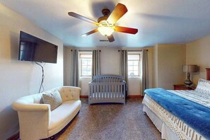 A bedroom with a ceiling fan, a television mounted on the wall, a crib, a bed with a blue blanket, and a small table with a lamp.