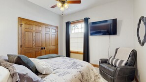Ein Schlafzimmer mit einem Bett, einem Sessel, einem Fernseher, einer Holztür und einem fenster mit Vorhängen.
