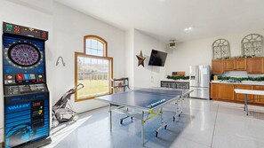 Ein Zimmer mit Tischtennisplatte, Dartscheibe, einer Stern-Dekoration, einem Fernseher und einer Küchenzeile mit Schränken und Kühlschrank.