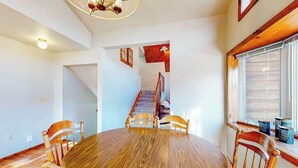 A dining area with a round wooden table and chairs, a staircase leading to another level, and a window with blinds. - Hilltop Getaway - Hilltop Home • Hot Tub & Billiards (Lead)