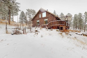 A wooden cabin with a deck and stairs leading to a second-story balcony, surrounded by snow-covered ground and trees.