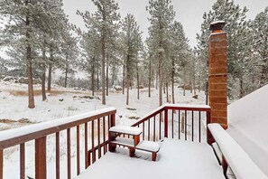 A wooden deck with a bench and a brick chimney, surrounded by snow-covered trees.